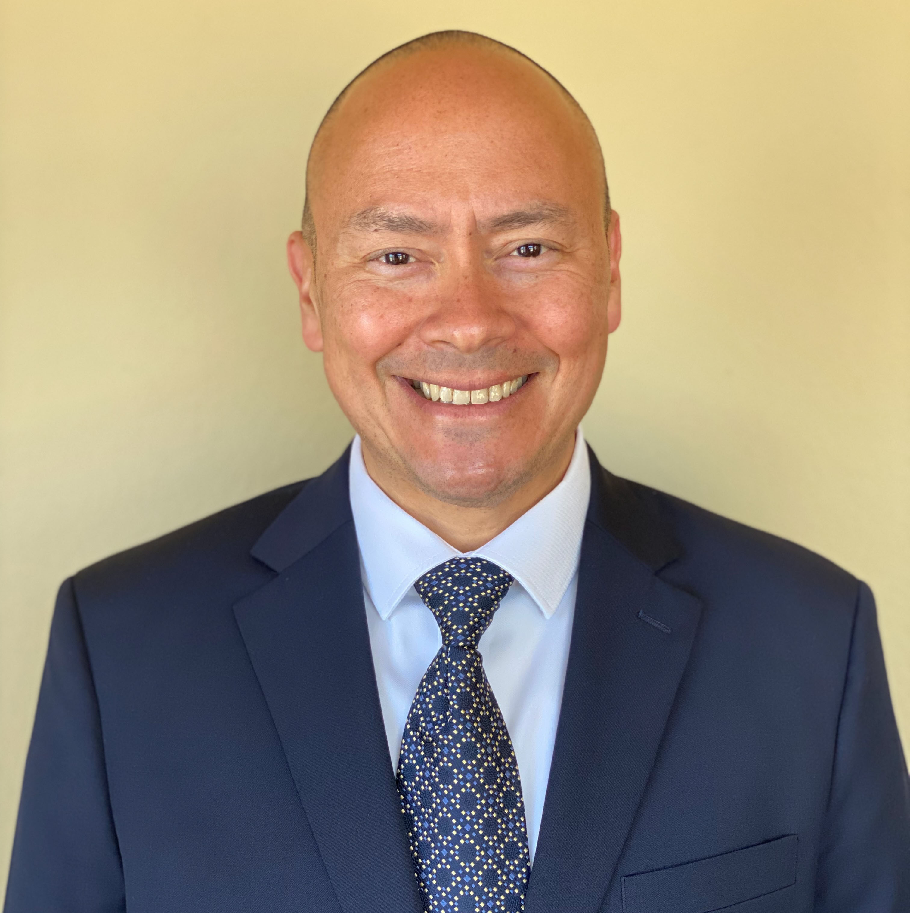 A head-and-shoulders portrait of a smiling man with a shaved head and light-to-medium skin tone. He is wearing a dark suit jacket, a light-colored dress shirt, and a patterned tie. The background is plain and softly lit in warm beige tones.